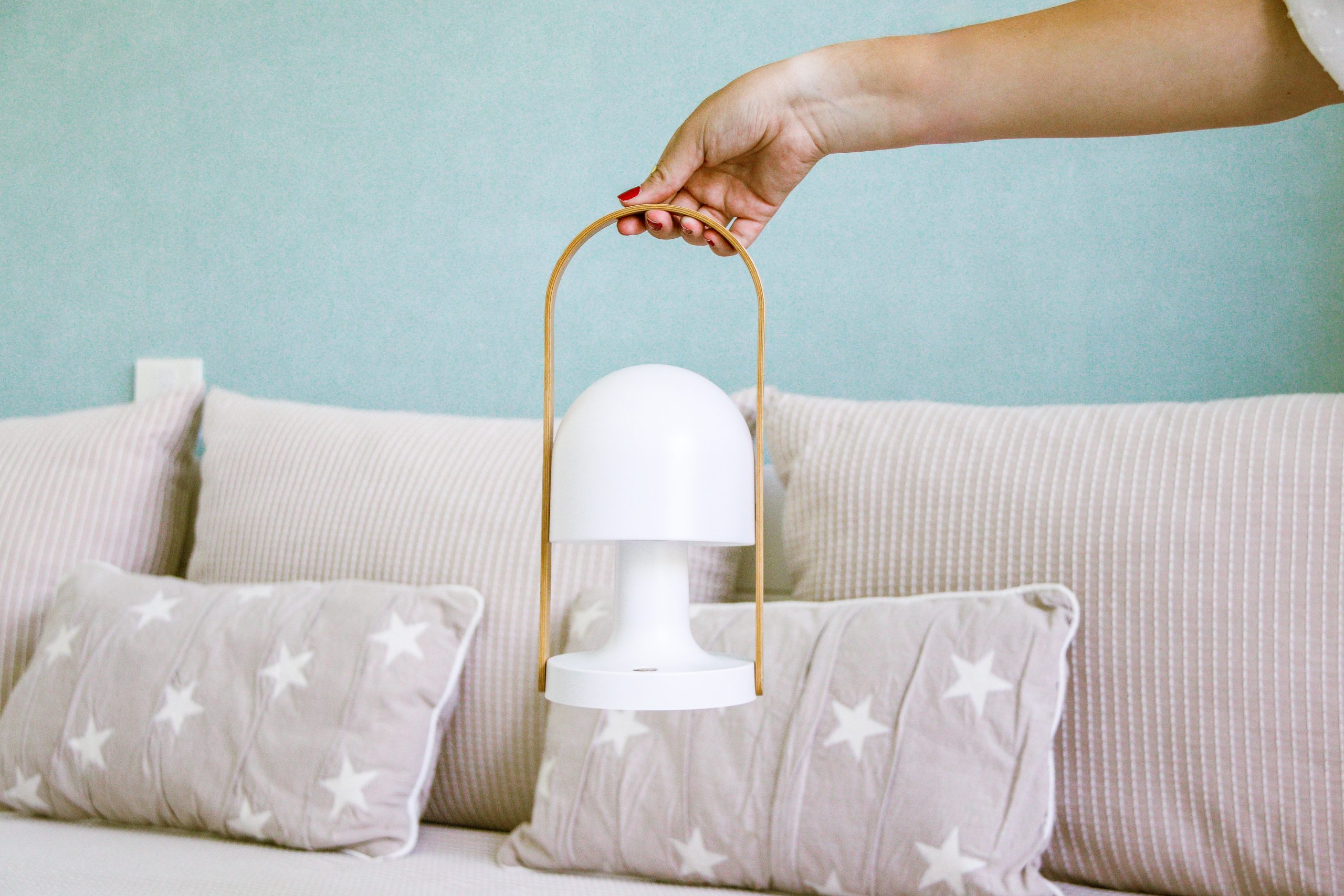 Fotografía profesional de una mano de mujer sujetando una lámpara de mesa. El asa que la sujeta es de madera y la lámpara blanca.